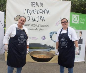 2 mujeres sonríen mientras sujetan un caldero con el guiso de la espadeñá