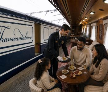 Tren Transcantábrico vista interior y exterior. En la imagen se muestra a una Familia sentada en mesa del tren de lujo Transcantábrico mientras un camarero sirve aperitivos, con el vagón visible desde el andén. 