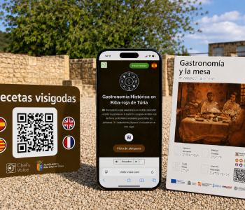Montaje de los recursos de Chef’s Voice instalados en el yacimiento arqueológico de Pla de Nadal. En primer plano se muestran una pegatina con código QR para acceder a las recetas visigodas, un teléfono móvil con la plataforma Chef’s Voice, donde se ha desarrollado la experiencia de Gastronomía Histórica de Riba-roja de Túria, y un panel tacto-visual con información sobre la gastronomía y la mesa, que incorpora braille, relieve y un código QR. Al fondo se observan los restos arqueológicos del yacimiento. 