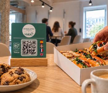Placa de Chef’s Voice con código QR y soporte de madera junto a sándwiches, cookies y café. Una mano coge un sándwich, con personas desenfocadas al fondo, en una recreación del catering para encuentros profesionales como los celebrados en Basque Living by HABIC. 