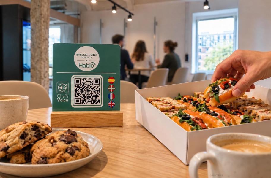 Placa de Chef’s Voice con código QR y soporte de madera junto a sándwiches, cookies y café. Una mano coge un sándwich, con personas desenfocadas al fondo, en una recreación del catering para encuentros profesionales como los celebrados en Basque Living by HABIC. 